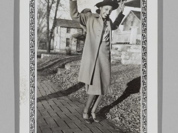 a woman in a coat and hat poses playfully on a sidewalk with her arms in the air