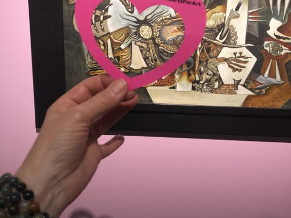 A hand holding a paper heart in front of the artwork, “L’ecartele (El descuartizado)” by Gunther Gerzso.