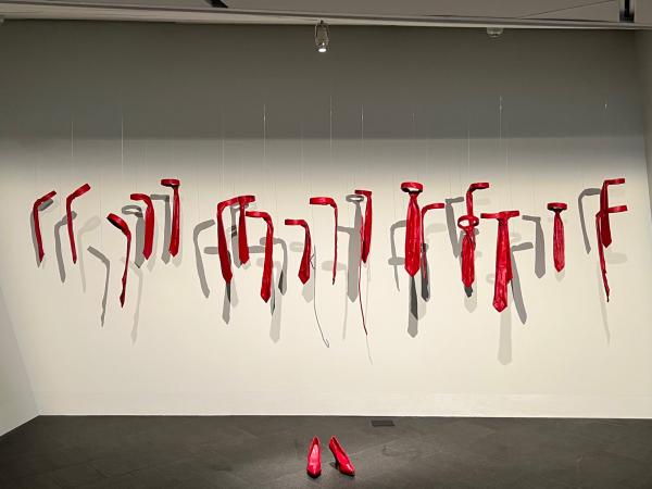 Pair of heels made out of lipstick wax on the ground, surrounded by suspended ties and an unfolded hanger, both also made out of lipstick wax. 