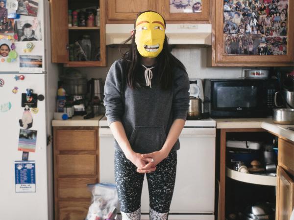 Young woman standing in messy kitchen wearing a yellow mask over her face