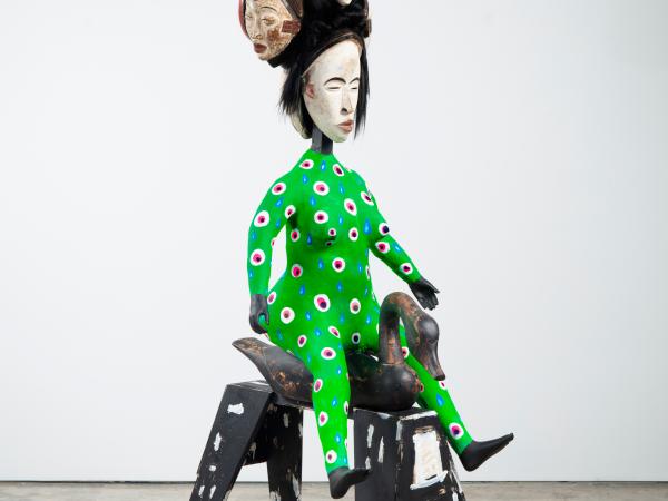 Otherworldly sculpture of a woman-like figure on a stool, with a headdress composed of several faces