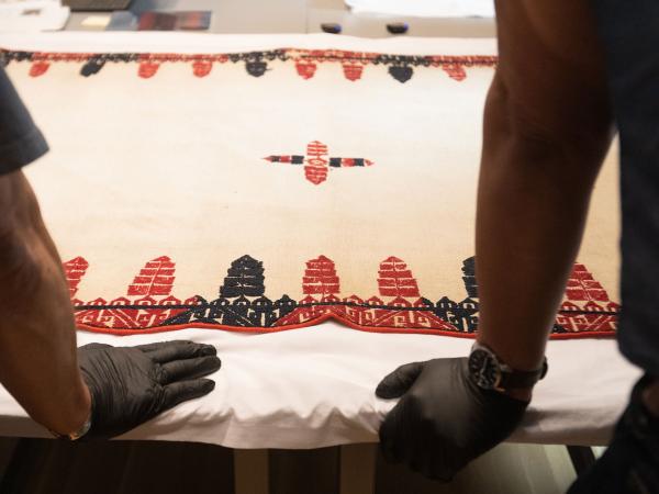 gloved hands on a table with a textile that is white with black and red designs embroidered on it; mannequins are in the background