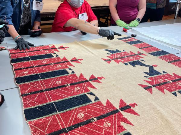people by a table with a textile on it. a man points at the textile