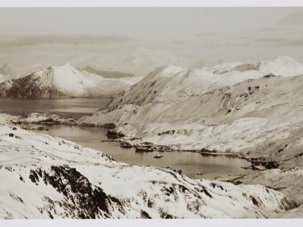 A sepia-like photograph of a bay with ships in the middle of a frozen hilly landscape.