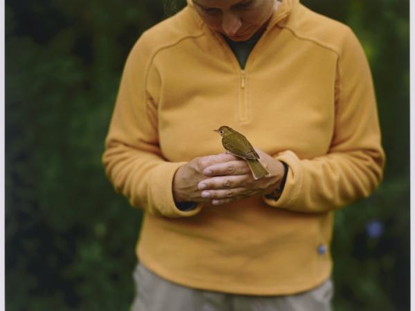 A woman in a light orange fleece jacket holds her hand together in front of her while facing the camera. A small yellow bird is perched on her hands facing the woman.