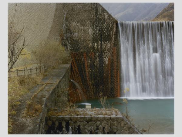 Water flows over a brickwork dam. Bright streaks of ruddy brown run vertically to the side of the waterfall, implying that water has run over that part of the dam wall in the past.