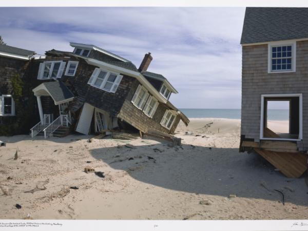A beach house crumbles on a sandy coastal beach, clearly the consequence of water flooding in the aftermath of a storm.