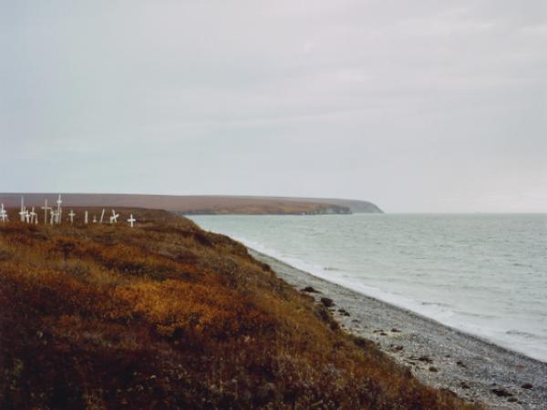 A photograph looking down a hilly coastline on a cloudy day. On one of the coastal hills in the middle distance, a collection of graveyard markers sit at various angles.