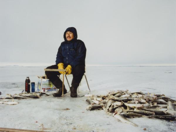 A man in winter outerwear sits in the middle of an icy lake on a stool by a pile of recently caught cod. A spear-like fishing tool sits on the ground between the figure and the camera.
