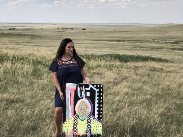 Danielle SeeWalker outside on the prairie standing next to her painting of Sitting Bull