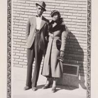 a man in a suit and hat and a woman in a coat in front of a brick wall