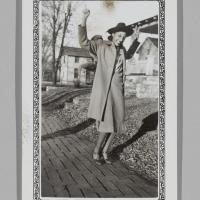 a woman in a coat and hat poses playfully on a sidewalk with her arms in the air