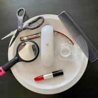 Objects arranged on a white plate and black background to look like a face. Objects include a lipstick container, a stapler for the nose, one eye created out of a mirrored hairbrush and another out of a tape dispenser. Two eyebrows are formed with hair barrettes, and on the edge of the plate are scissors and a comb