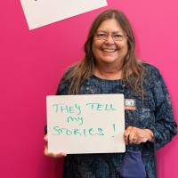 Woman posing with a written sign that says "I take photos because they tell my stories!"