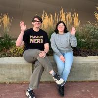 Shop employee wearing shirt that says Museum Nerd sits next to another employee on a bench outside the museum