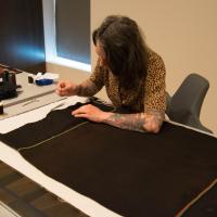 conservator at a table sewing a long brown textile