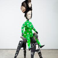 Otherworldly sculpture of a woman-like figure on a stool, with a headdress composed of several faces