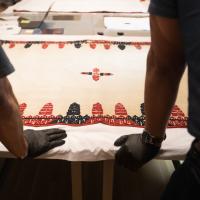 gloved hands on a table with a textile that is white with black and red designs embroidered on it; mannequins are in the background