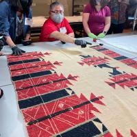 people by a table with a textile on it. a man points at the textile