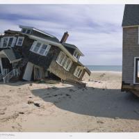 A beach house crumbles on a sandy coastal beach, clearly the consequence of water flooding in the aftermath of a storm.