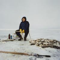 A man in winter outerwear sits in the middle of an icy lake on a stool by a pile of recently caught cod. A spear-like fishing tool sits on the ground between the figure and the camera.