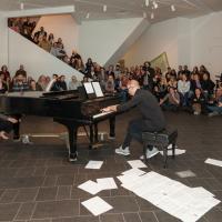 musician at a grand piano surrounded by papers on the floor and a large crowd at Untitled Final Friday