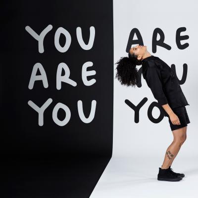 Shantell Martin in front of a wall with her signature drawings of the words You are you are you you