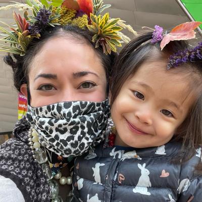 a woman wearing a facemask and a flower crown cheek to cheek with young daughter wearing flowers in her hair