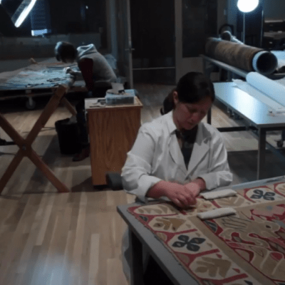 Video still taken from a series of videos documenting the conservation of a Spanish Colonial table cover at the Denver Art Museum. This image shows a textile conservator repairing the tapestry.