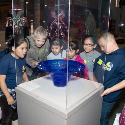 Children and adult standing around looking closely at the blue glass vase ( in the center