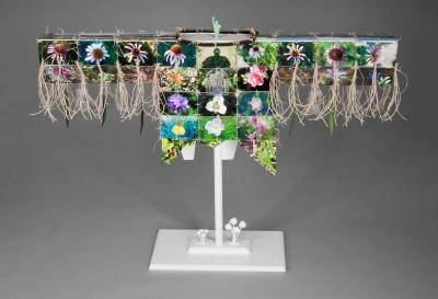 A war shirt made out of found objects like glass beads, velvet, silk plant leaves, rubber figures, color photographs, an Ethernet cord, a SD card, plastic packaging, and an hemp cord.