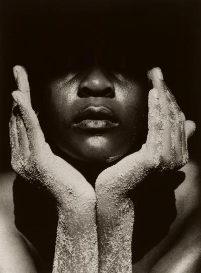 Close-up of a Black person with their face in their hands; their hands are covered in sand and their eyes, and the top of their head is shrouded in darkness.