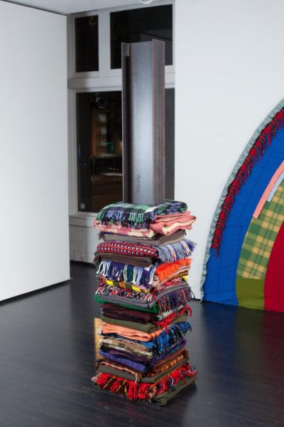 Dozens of colorful blankets folded and stacked vertically. A gray steel beam comes out from the top of the stack and extends upwards.