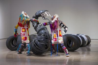 Large-scale sculptural installation made up of oil drums, ammunition cans, trash, and found objects arranged in a snake-like pattern.