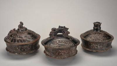 Three lidded bowls that are graduated in size. The bowl on the left features water lilies and hummingbirds, the middle bowl has a fish-shaped handle and reptilian imagery, and the bowl on the right depicts a deity riding on a mythical bird.