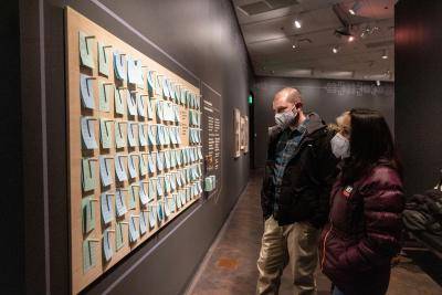 Masked man and woman looking at a wall of notes and responses written by other museumgoers.