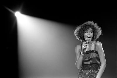 black and white photo of Whitney Houston under a spotlight, singing into a microphone