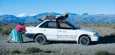 Woman pushing a car with a man sprawled on his back on the roof, against a mountain backdrop