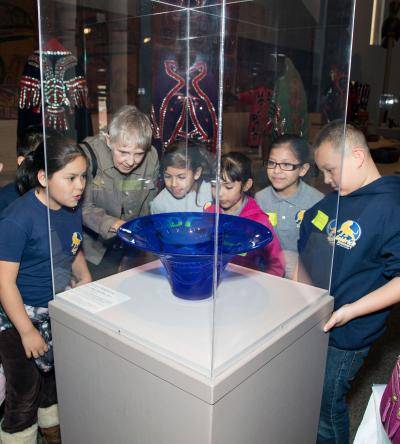 Children and adult standing around looking closely at the blue glass vase ( in the center