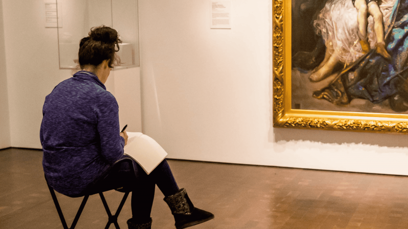 Person sitting in a gallery at the Denver Art Museum writing in a journal