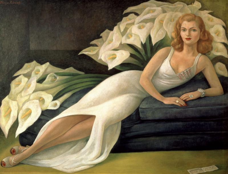 Natasha Gelman lying on a sofa in a long white gown reminiscent of the large white calla lillies behind her