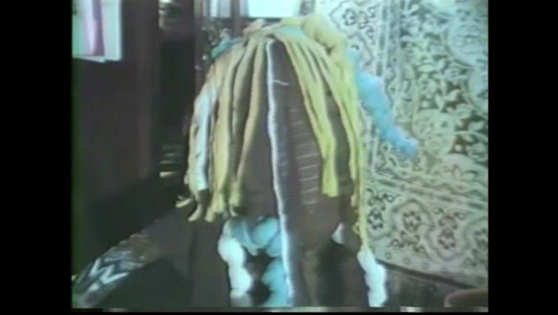 Still image from a video of Nengudi in the process of removing a mask made of long pieces of multicolored nylon pantyhose.