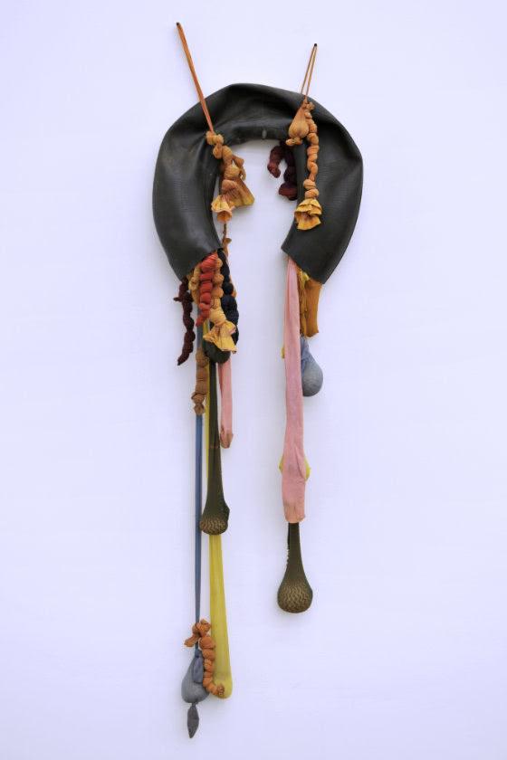 Sculpture hanging on wall featuring upside down U-shaped black rubber tube with multiple nylon pantyhose hanging from each end. The pantyhose are knotted and weighted with sand.
