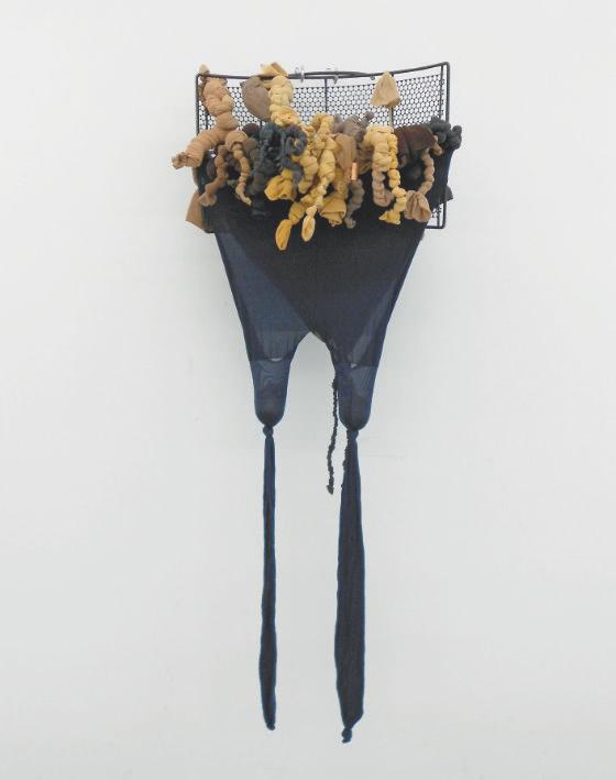 Attached to a square wire grid on the wall, a pair of navy blue nylon pantyhose hang down in two long strips. At the top are knotted pieces of yellow, brown and blue nylon.