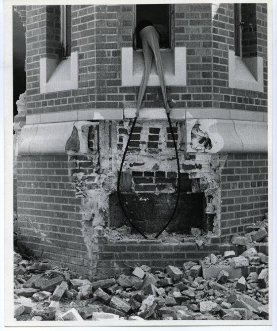 Black and white photograph of Nengudi leaning out the window of a crumbling building. She wears pantyhose on her head attached to two long pieces of wire.