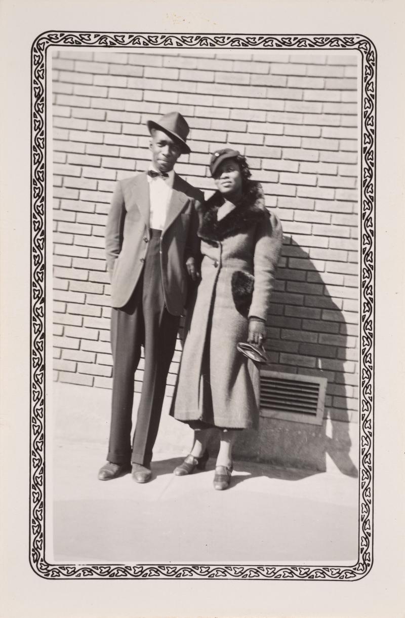 a man in a suit and hat and a woman in a coat in front of a brick wall