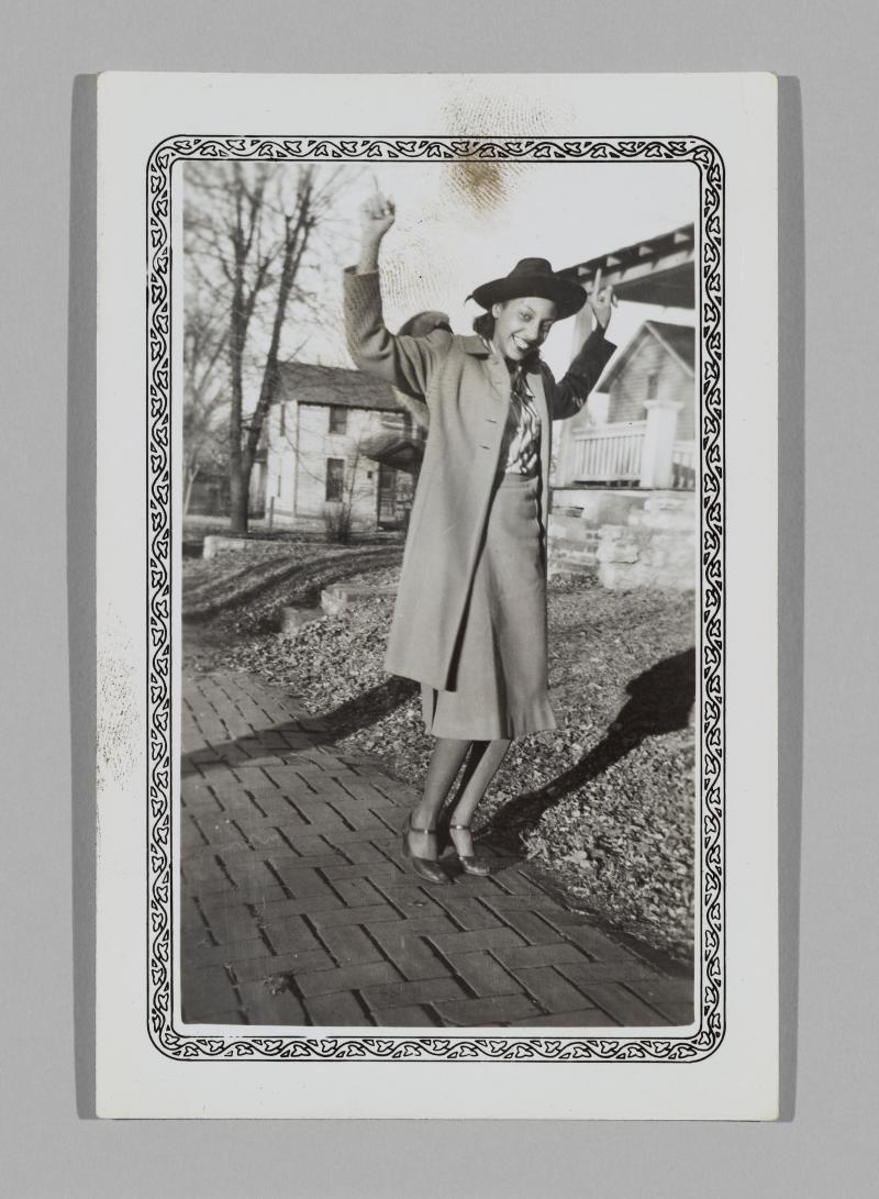a woman in a coat and hat poses playfully on a sidewalk with her arms in the air