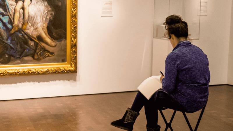 Person sitting in a gallery at the Denver Art Museum writing in a journal.