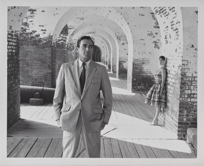 Black and white photograph of couple under an arched walkway. Gregory in a suit and tie in the foreground and Véronique in a dress leaning against a brick arch in the background.