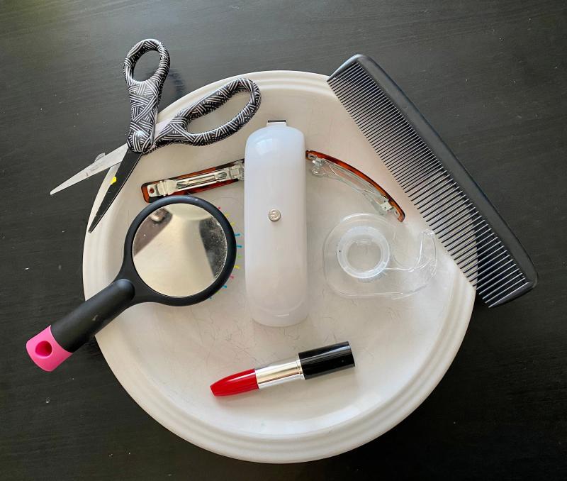 Objects arranged on a white plate and black background to look like a face. Objects include a lipstick container, a stapler for the nose, one eye created out of a mirrored hairbrush and another out of a tape dispenser. Two eyebrows are formed with hair barrettes, and on the edge of the plate are scissors and a comb